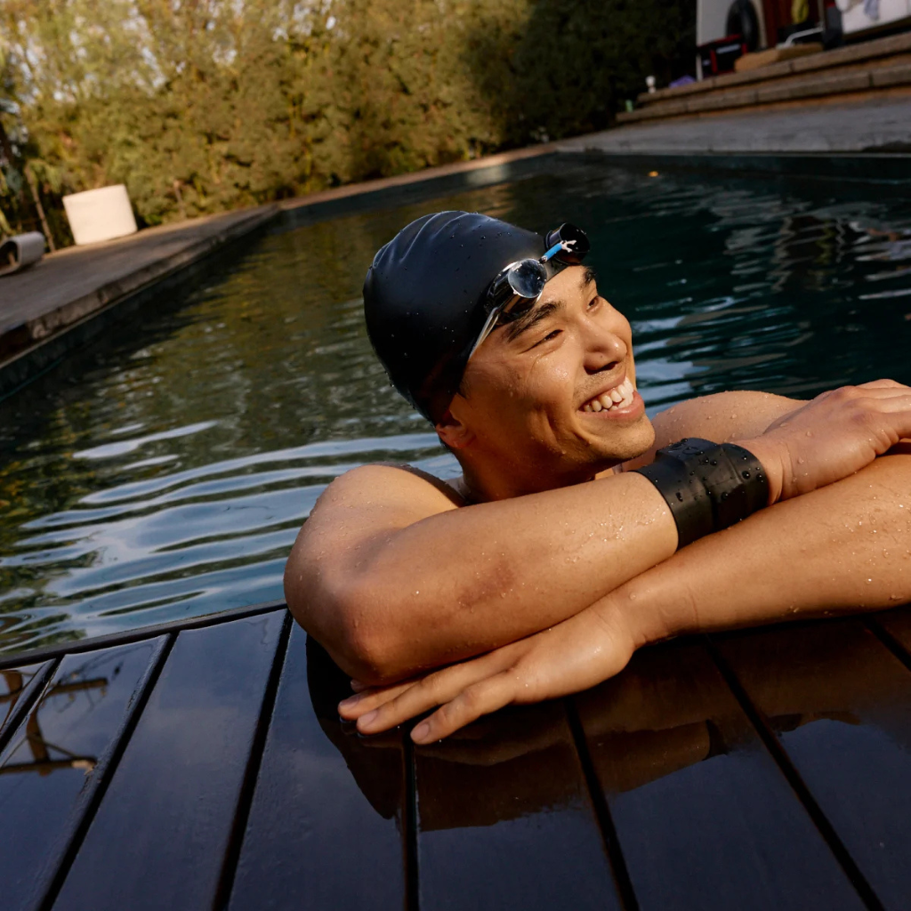 Person wearing a swim cap and goggles, and WHOOP MG leaning on a poolside ledge - Bikers.SG