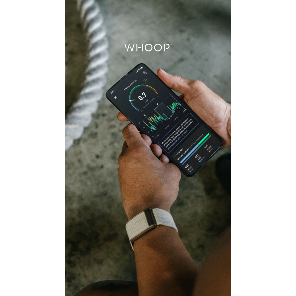 A person holding a smartphone showing the WHOOP fitness app screen, with a white WHOOP Peak 5.0 on their wrist. The background is a textured gray concrete floor with a white rope - Bikers.SG